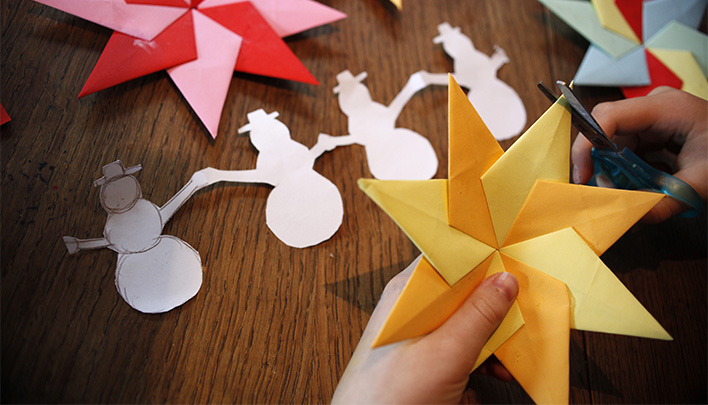 Eine Hand hält einen Weihnachtsstern aus orangenem und gelbem Papier. Im Hintergrund auf einem braunen Tisch weitere Weihnachtssterne und eine Schneemanngirlande.