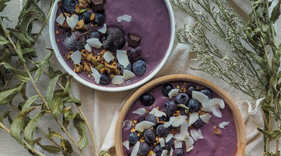 lifestyle-flatlay-zwei-schalen-mit-acaipueree-blaubeeren-kokoschips-weisse-tischdecke-getrocknete-kraeuter-04-24-96381-0