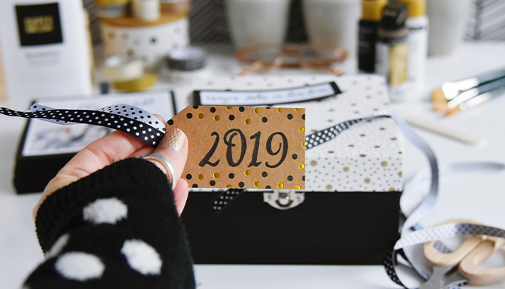 Eine Hand hält einen Geschenkanhänger aus braunem Karton mit schwarzer Schrift und dem Jahr 2019. Im Hintergrund die fertige Leporello Foto-Box