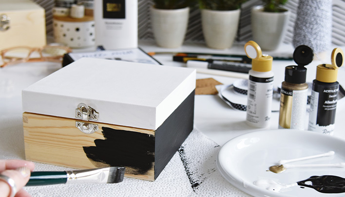 Eine hellbraune Holzbox mit weiß angemaltem Deckel wird schwarz angemalt