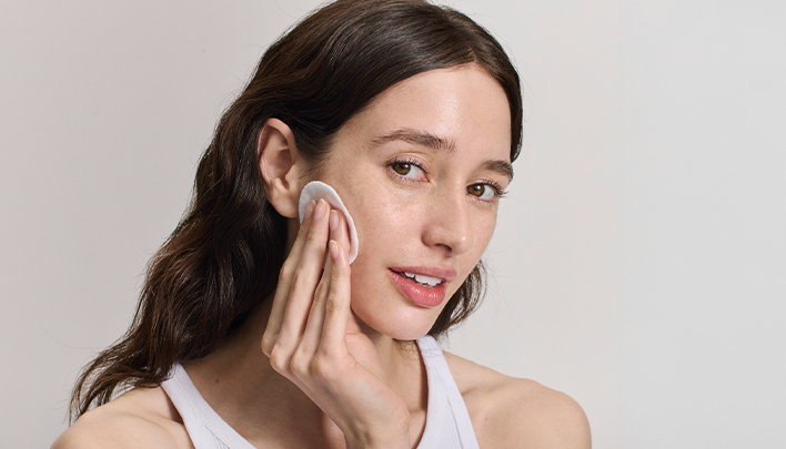 Eine dunkelhaarige Frau mit offenen Haaren in Nahaufnahme tupft sich das Gesicht mit einem Wattepad ab