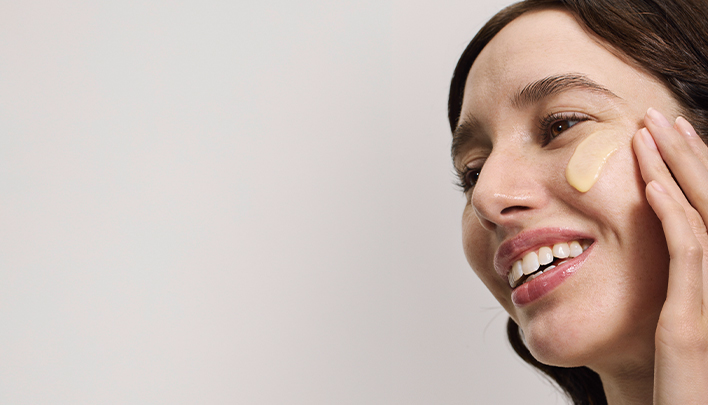 Eine dunkelhaarige Frau in Nahaufnahme trägt sich Creme auf die Wange auf