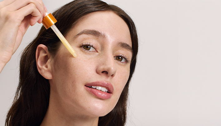 Eine dunkelhaarige Frau in Nahaufnahme und offenen Haaren tupft sich mit einer Pipette eine Flüssigkeit unter die Augen