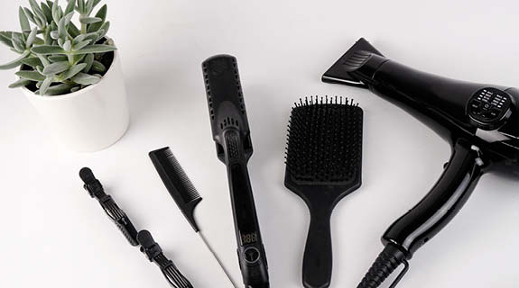 lifestyle-fuenf-schwarze-friseurtools-haarspangen-kamm-glaetteisen-buerste-foen-gruene-pflanze-weisser-hintergrund-575x320-90676-0