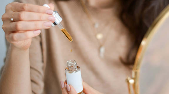 lifestyle-brunette-frau-haelt-geoeffnetes-pipettenflaeschchen-mit-goldfarbener-fluessigkeit-575x320-91991-0