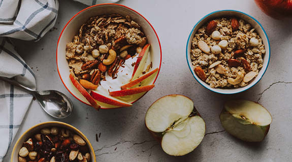 lifestyle-drei-schalen-mit-muesli-und-nuessen-angeschnittener-apfel-silberner-loeffel-kariertes-geschirrtuch-weiss-grauer-hintergrund-575x320-90548-0