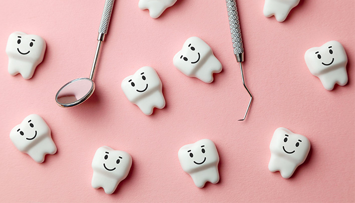Modell-Zähne mit lachenden Gesichtern auf rosa Hintergrund, dazwischen ein Mundspiegel und ein Zahnschaber