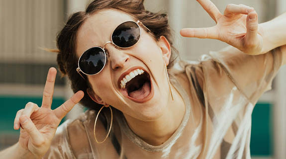 lifestyle-junge-frau-sonnenbrille-peacezeichen-offenes-lachen-zaehne-04-24-96464-0