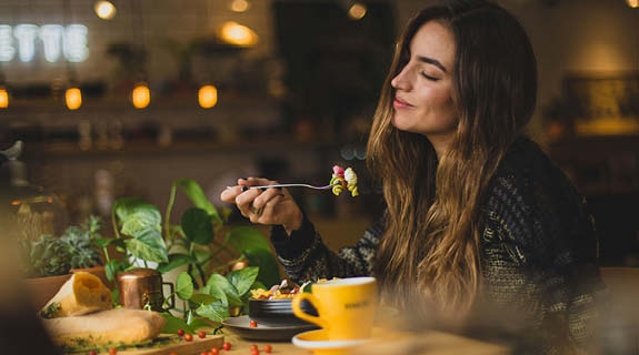 lifestyle-brunette-junge-frau-am-tisch-beim-essen-pflanzen-gelbe-tasse-innenraum-im-hintergrund-575x320-92299-0