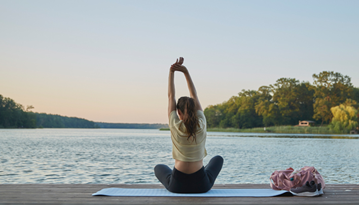Person macht Yoga am See und streckt die Arme im Morgenlicht