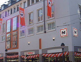 Vonkajší pohľad na modernizovanú predajňu Müller z 2000. rokov so zdobeným vstupom a vlajkami.