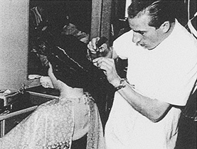 Historická fotografia kaderníka pracujúceho na účese zákazníčky, pripomínajúca začiatky spoločnosti Müller.